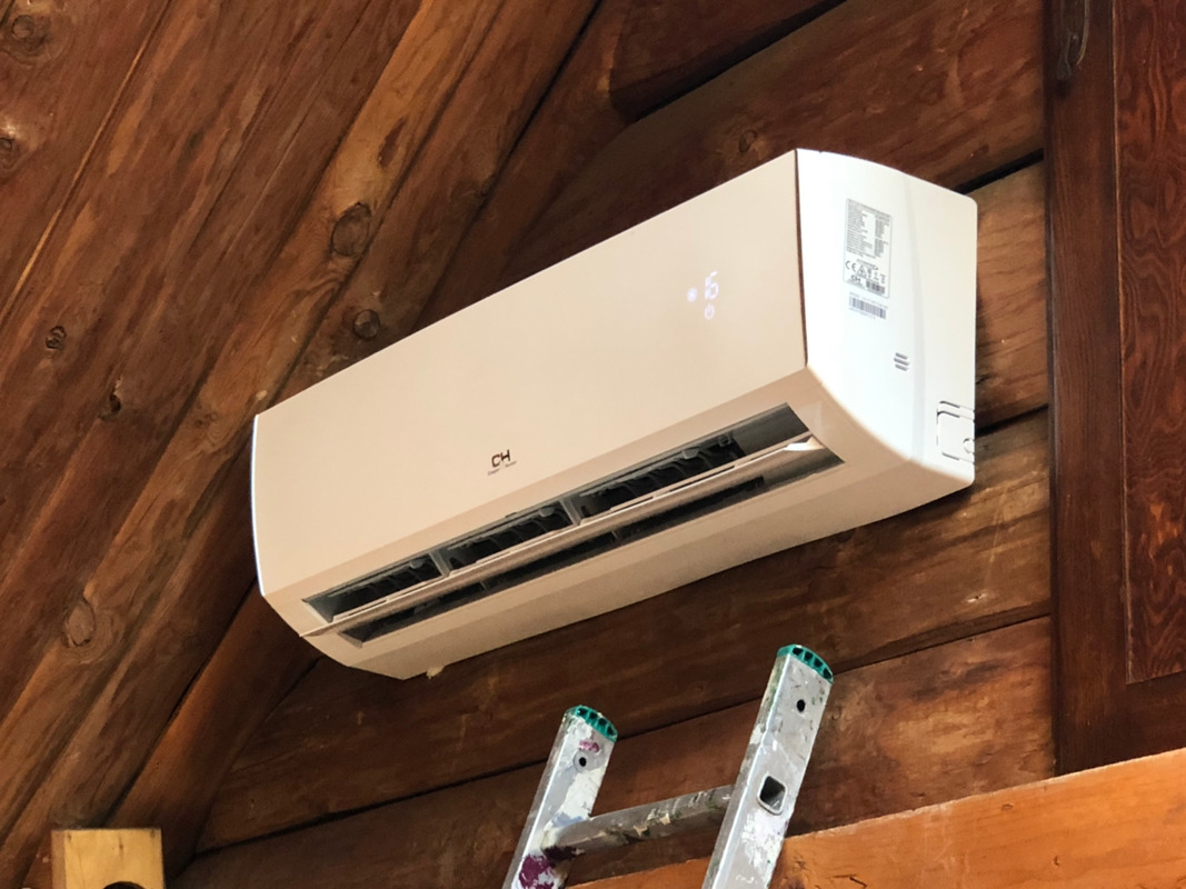 Biała jednostka klimatyzacji marki CH zamontowana na ścianie z drewnianych desek, widoczna drabina malarska.