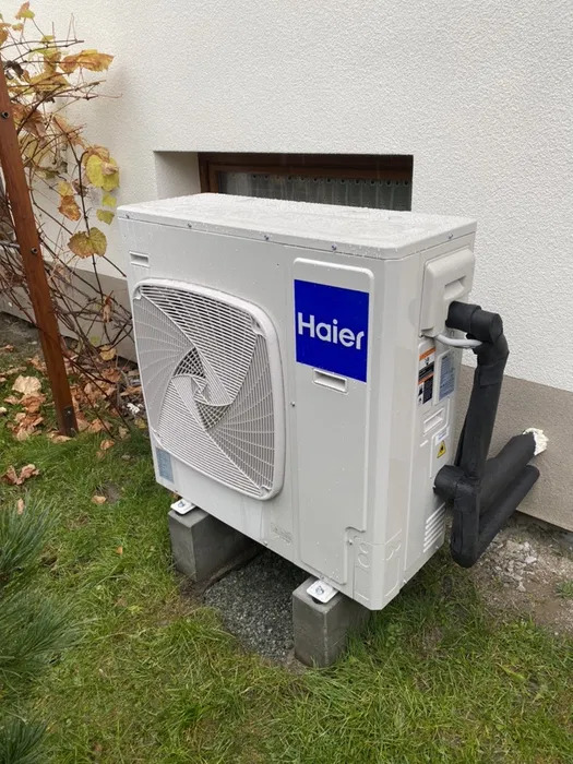 Biała jednostka zewnętrzna z niebieskim logo Haier na betonowych podstawach, widoczne rury w izolacji, trawa i krzew z żółtymi liśćmi w tle.