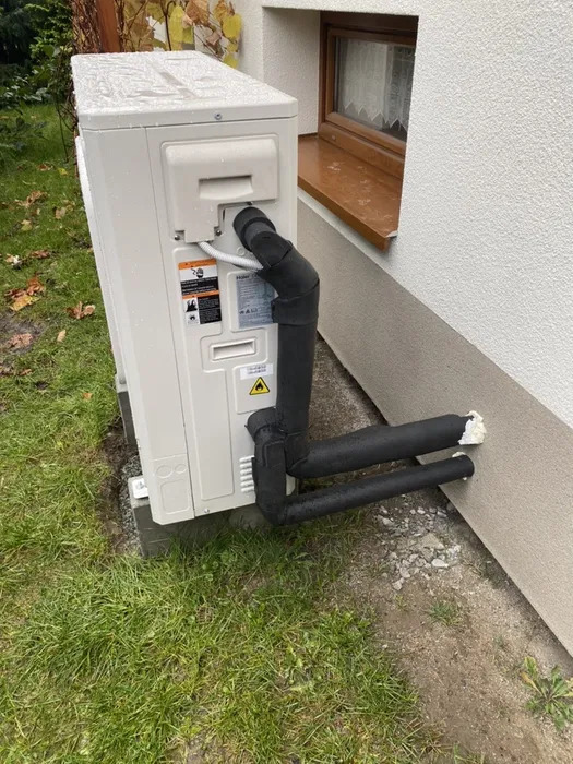 Zewnętrzna jednostka pompy ciepła zamontowana przy ścianie domu, widoczne ocieplone rury i podstawa betonowa na trawniku, lekki deszcz.