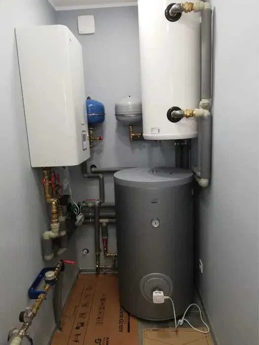 Kompaktowa kotłownia w pomieszczeniu: biały wiszący piec, pionowy zbiornik ciepłej wody i szary akumulacyjny zbiornik buforowy na podłodze, połączone rurami.