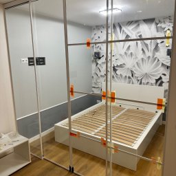 Iconglass Piotr Brejwo - Montaż szklanej ścianki działowej w sypialni, widoczne tymczasowe mocowania taśmą, w tle białe łóżko i ściana z motywem roślinnym.