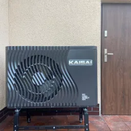 Zewnętrzna jednostka pompy ciepła Kaisaí na metalowym stelażu, zainstalowana przy beżowej ścianie i brązowych drzwiach, widoczna z bliska na brązowej terakocie.