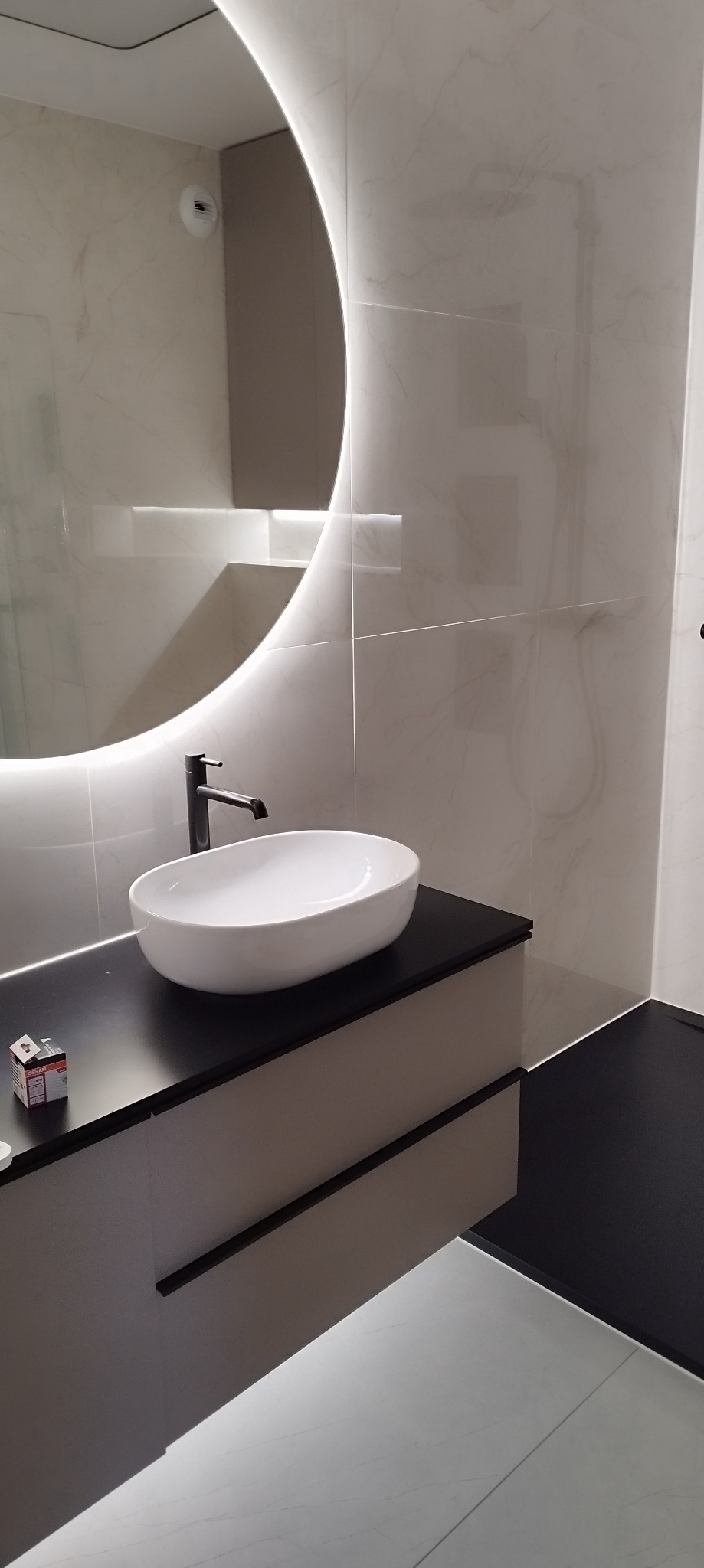 Nowoczesna łazienka z okrągłym lustrem podświetlanym LED, białą umywalką nablatową i czarną armaturą oraz blatem, marmurowe płytki ścienne i czarna podłoga.