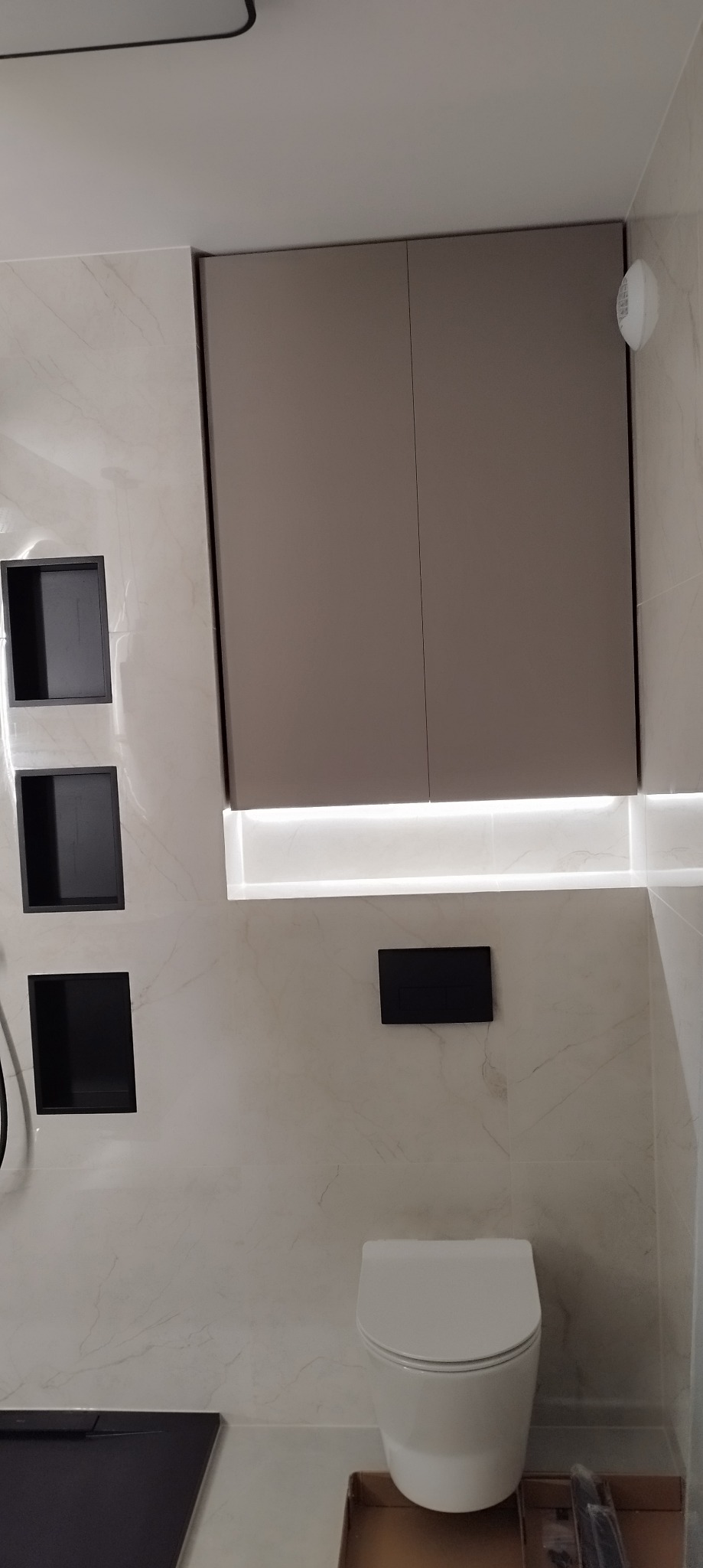 Nowoczesna łazienka z podświetlanymi wnękami w marmurowej ścianie, wiszącą szafką w kolorze taupe i minimalistyczną toaletą.