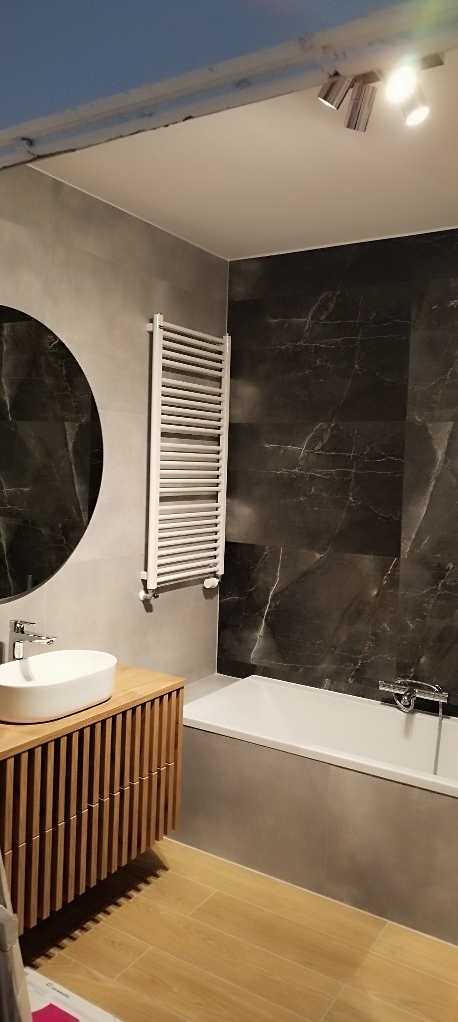 Nowoczesna łazienka z umywalką nablatową, szafką z lamelami, grzejnikiem drabinkowym i czarną płytką imitującą marmur na ścianie, oświetlona punktowo.