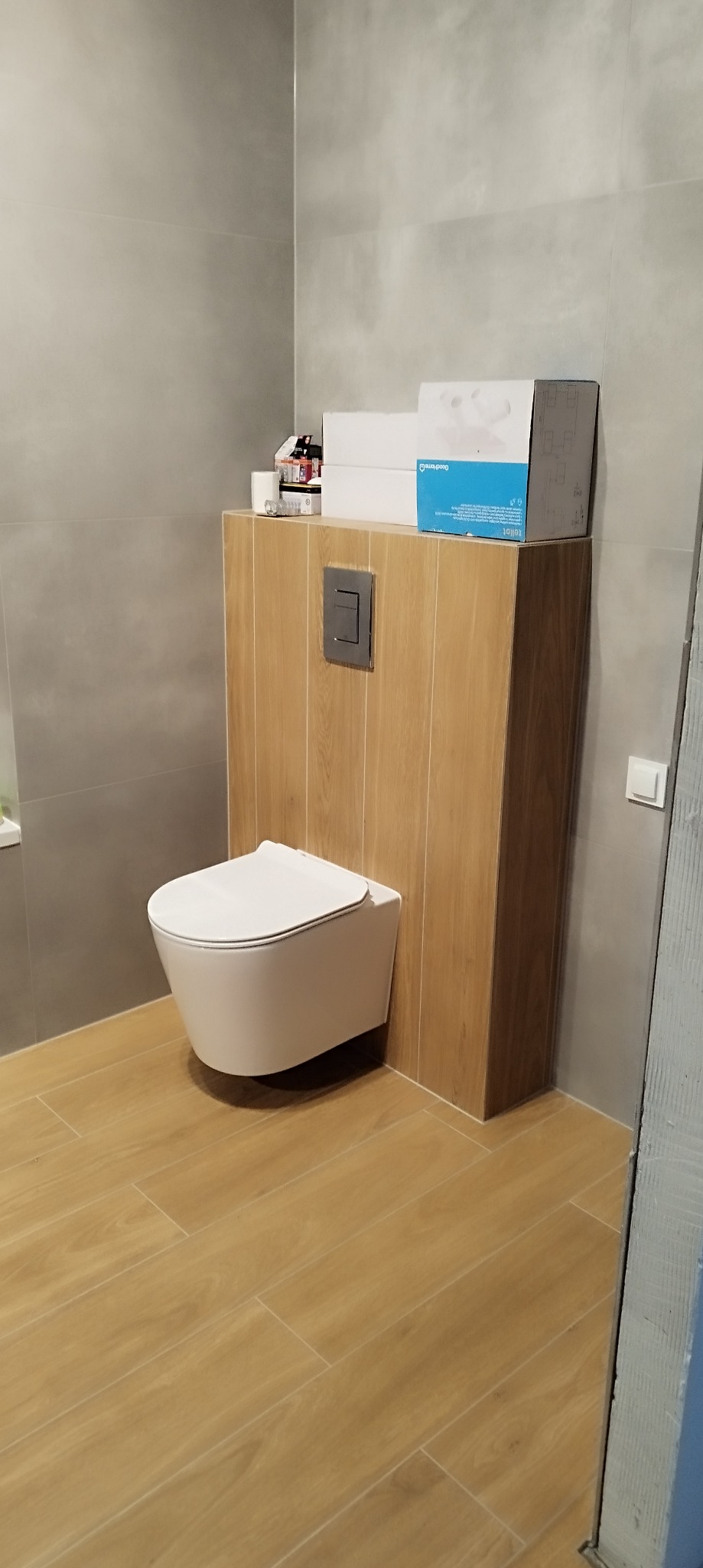 Nowoczesna, podwieszana toaleta z zabudową z drewnopodobnych paneli, szare płytki ścienne i podłogowe, pudełka i kosmetyki na zabudowie.