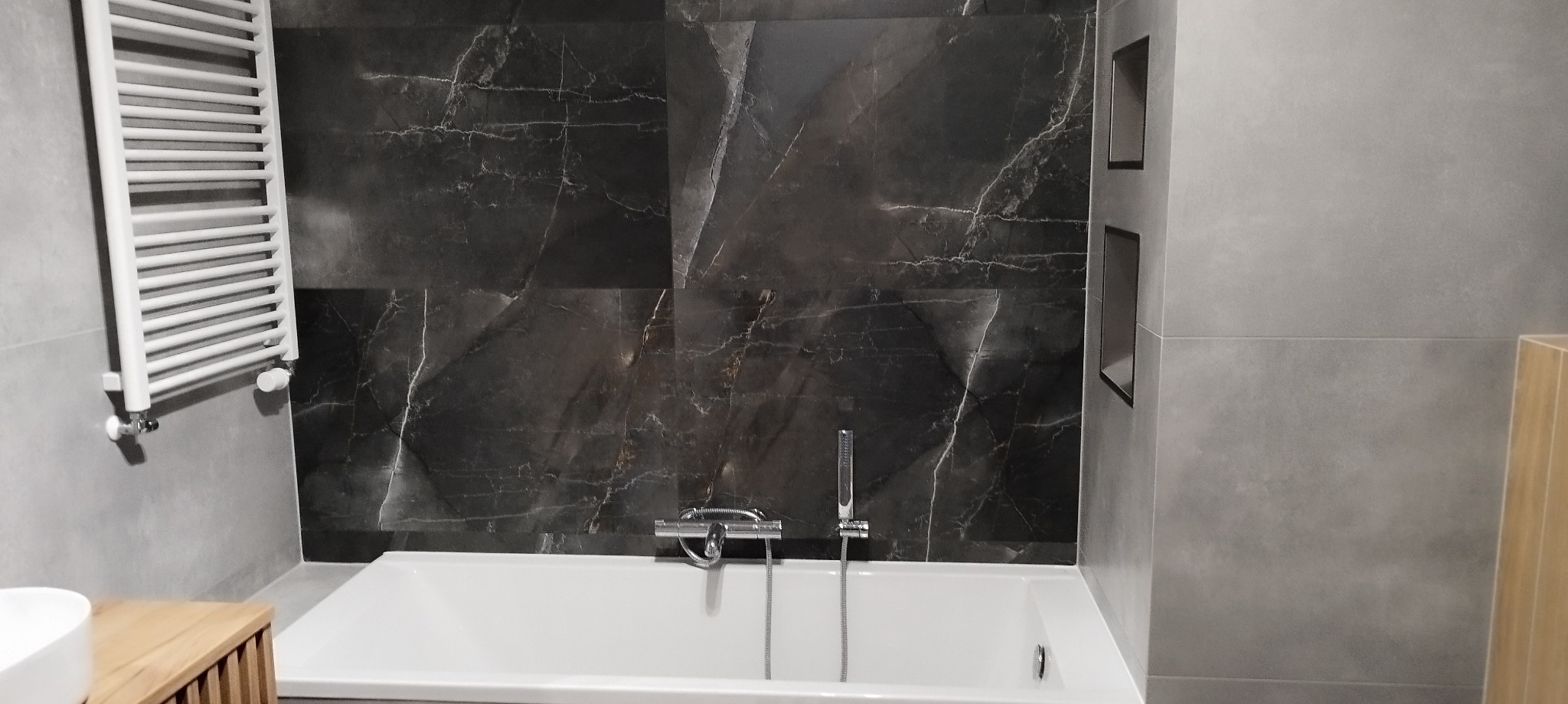Nowoczesna łazienka z wanną, ciemnymi płytkami imitującymi marmur na ścianie oraz grzejnikiem drabinkowym po lewej stronie.