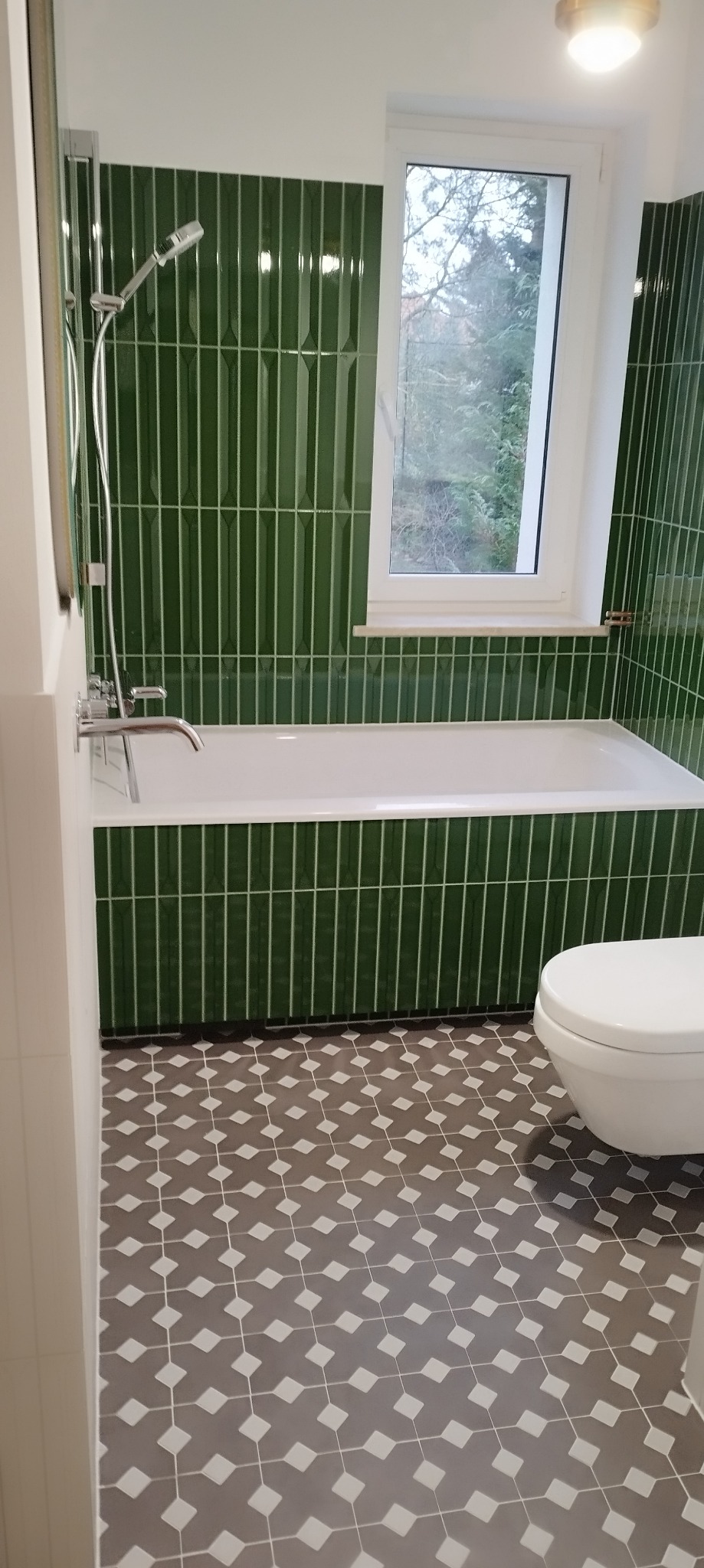 Nowoczesna łazienka z wanną i toaletą, wykończona pionowymi, butelkowozielonymi płytkami i geometryczną podłogą w odcieniach szarości.