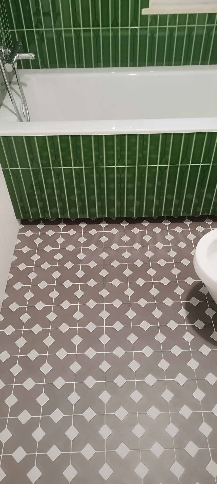 Łazienka z wanną obłożoną pionowymi, zielonymi płytkami oraz podłogą z szarych płytek w geometryczny wzór z białymi rombami.