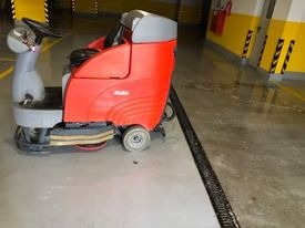 Czerwona maszyna czyszcząca podłogi z siedzeniem operatora na mokrej posadzce w garażu podziemnym, widoczne żółto-czarne pasy na filarach.