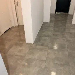 Ułożona szara, matowa terakota w korytarzu z białymi ścianami i drzwiami, widoczne fugi, odbicie światła na powierzchni płytek.