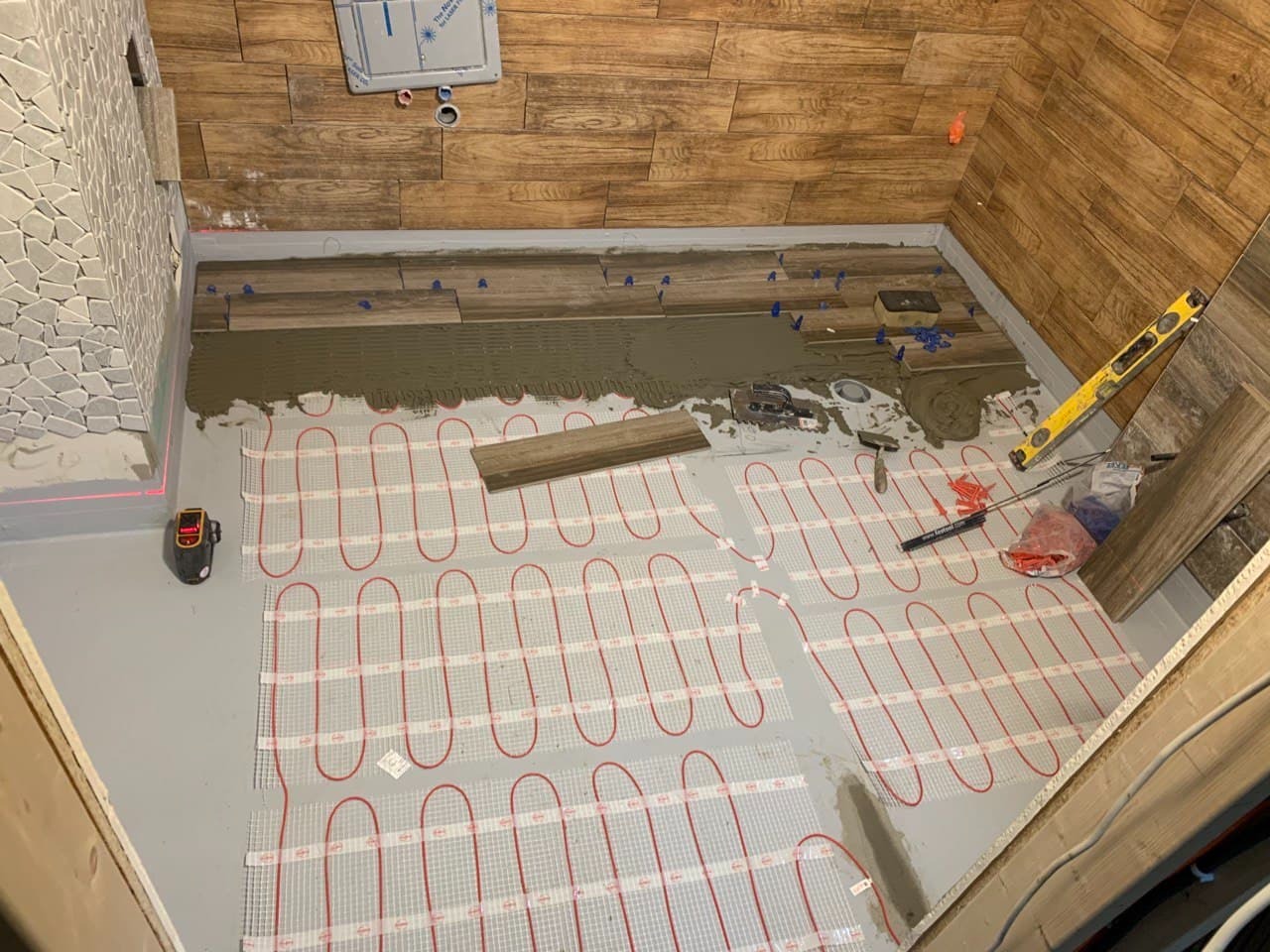Układanie płytek imitujących drewno na podłodze z ogrzewaniem podłogowym w trakcie remontu łazienki, widoczne narzędzia glazurnicze i poziomica.
