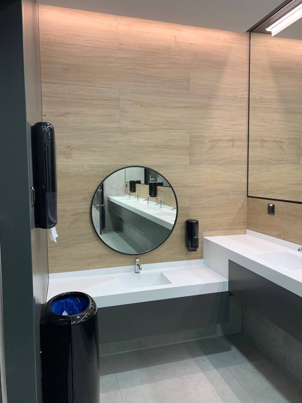 Nowoczesna łazienka publiczna z umywalkami w zabudowie, okrągłym lustrem i dozownikami, wykończona płytkami imitującymi drewno.