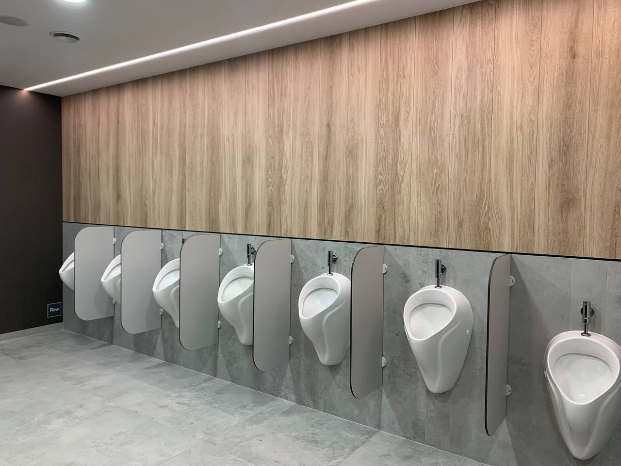 Publiczna toaleta męska w Ustroniu z rzędem białych pisuarów oddzielonych szarymi przegrodami, podświetlona listwą LED i wykończona drewnopodobną okładziną ścienną.