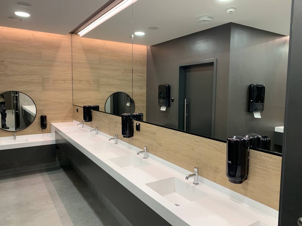 Nowoczesna łazienka publiczna z długim blatem z umywalkami, dużym lustrem, drewnopodobnymi panelami na ścianie i czarnymi dozownikami mydła i ręczników papierowych.