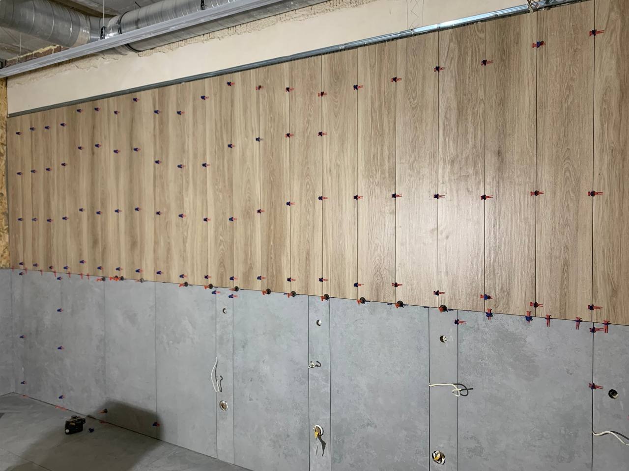 Układanie płytek imitujących drewno na ścianie, z widocznymi klipsami poziomującymi i szarymi płytkami poniżej, w trakcie prac wykończeniowych.