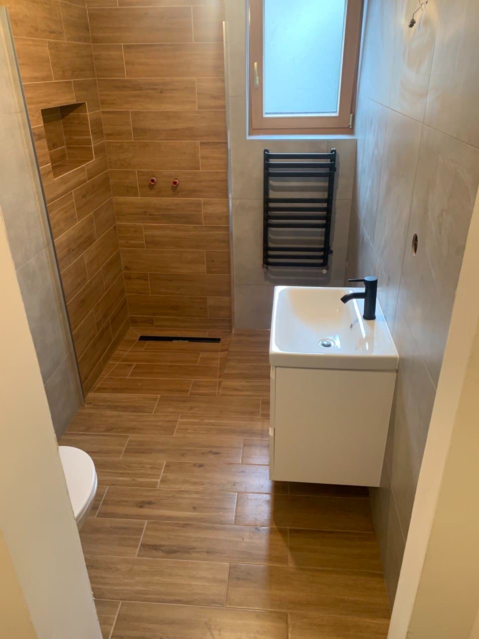 Nowoczesna łazienka z drewnopodobnymi płytkami, kabiną prysznicową typu walk-in, białą szafką z umywalką i czarnym grzejnikiem drabinkowym pod oknem.