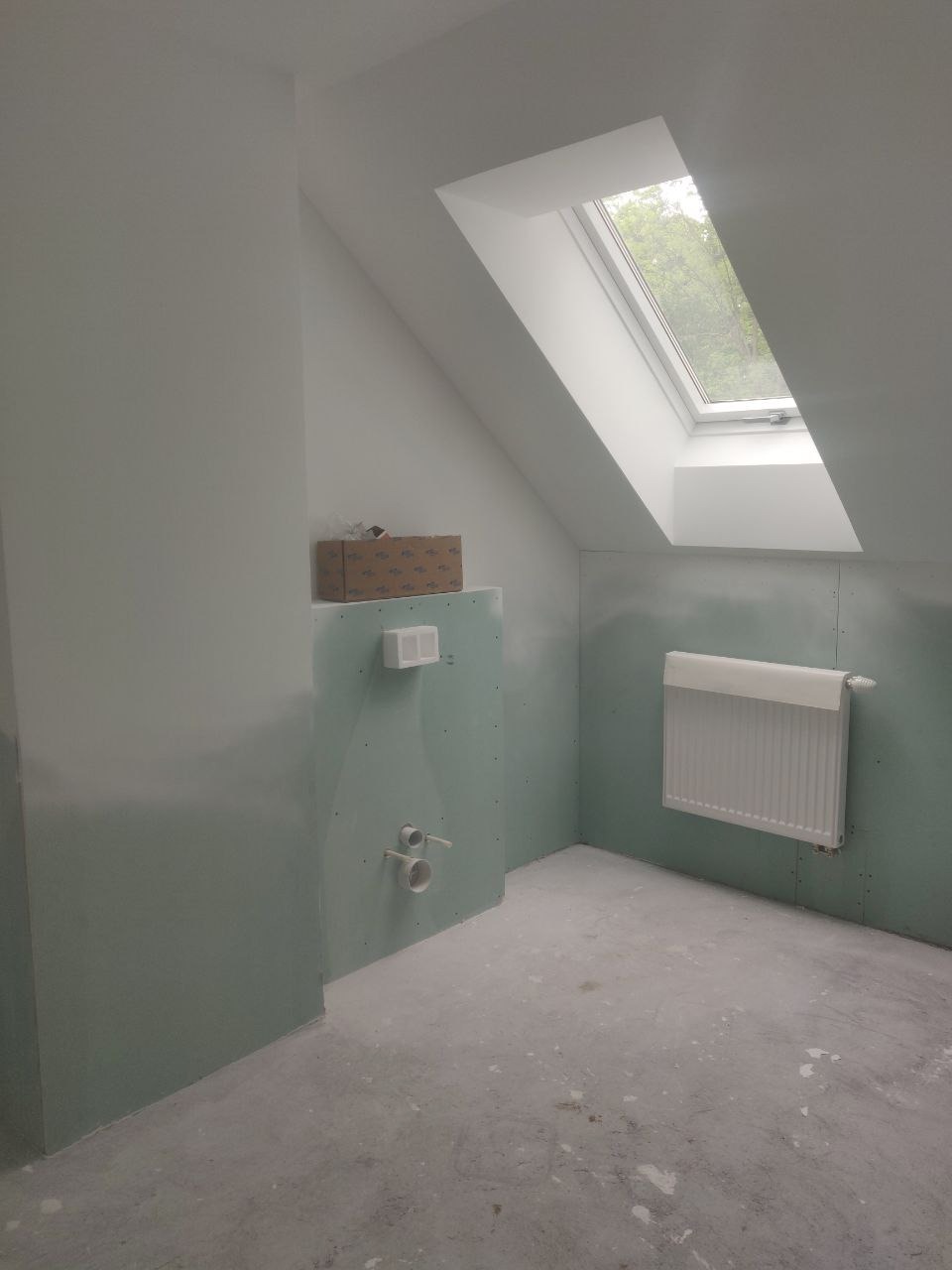 Łazienka na poddaszu w trakcie remontu: ściany wyłożone płytami gipsowo-kartonowymi, zamontowany grzejnik i instalacja wodno-kanalizacyjna, okno dachowe wpuszczające naturalne światło, karton...