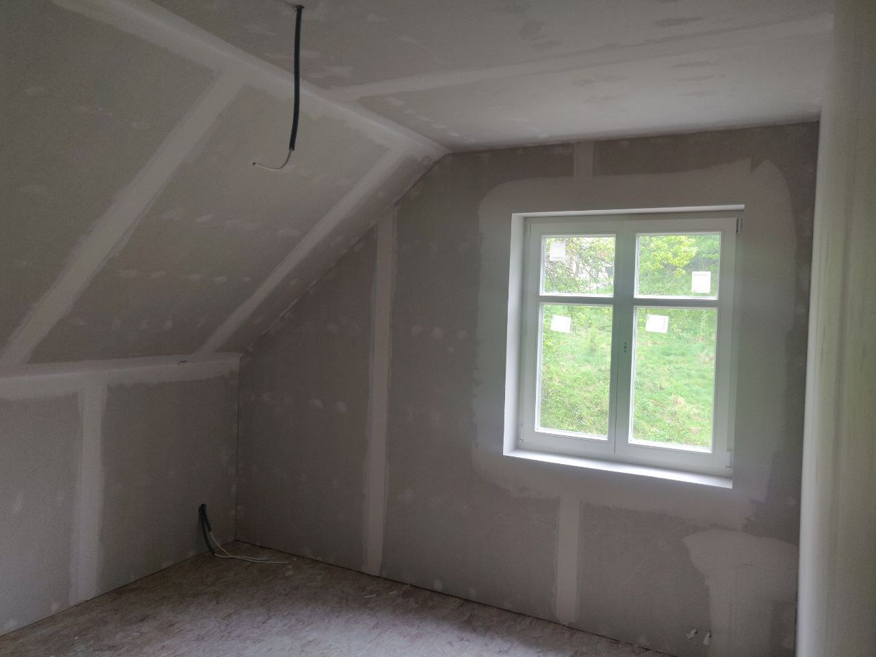 Poddasze w trakcie prac wykończeniowych: ściany i skosy obłożone płytami gipsowo-kartonowymi, widoczne łączenia wymagające szpachlowania, białe okno z widokiem na zieleń za nim, wystające przewody...