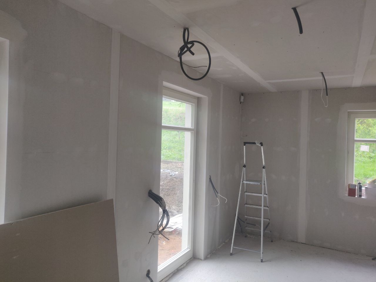 Pokój w trakcie prac wykończeniowych: ściany przygotowane do malowania, widoczne kable elektryczne, drabina i płyta gipsowo-kartonowa.
