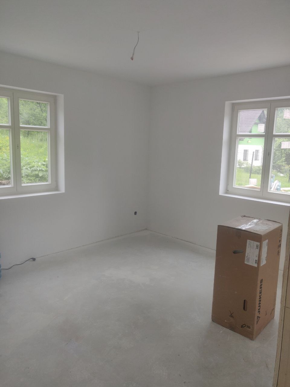 Wnętrze pokoju po remoncie, świeżo pomalowane ściany na biało, dwa okna z widokiem na zieleń, na podłodze wylewka, w rogu stoi karton firmy Junkers.