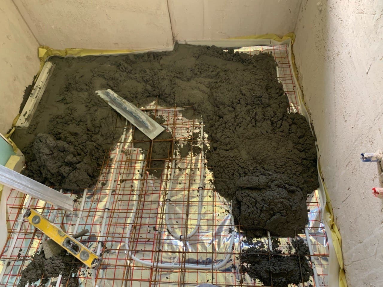 Wylewka betonowa na ogrzewaniu podłogowym w trakcie prac, widoczna poziomica i kielnia, ściany przygotowane do dalszych prac wykończeniowych.