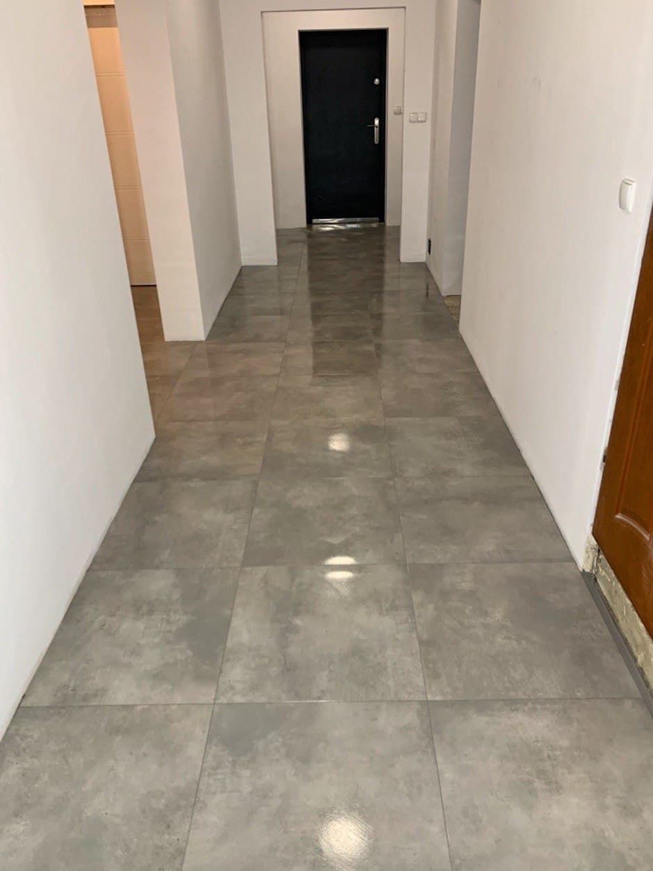 Wykończony korytarz z szarymi, kwadratowymi płytkami podłogowymi, białe ściany, widoczne drzwi wejściowe i fragment ościeżnicy.