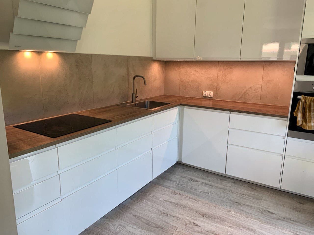 Nowoczesna kuchnia z białymi, błyszczącymi szafkami, drewnianym blatem i płytą indukcyjną, oświetlona punktowo pod szafkami wiszącymi, z szarą, teksturowaną ścianą nad blatem i drewnopodobną podłogą.