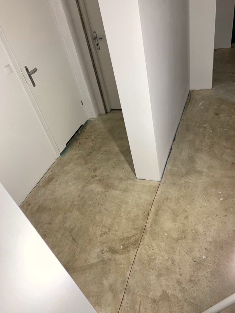 Przygotowanie do remontu: surowy beton na podłodze, świeżo pomalowane białe ściany i drzwi w tle, widoczny fragment niebieskiej taśmy malarskiej zabezpieczającej podłogę przy ścianie.