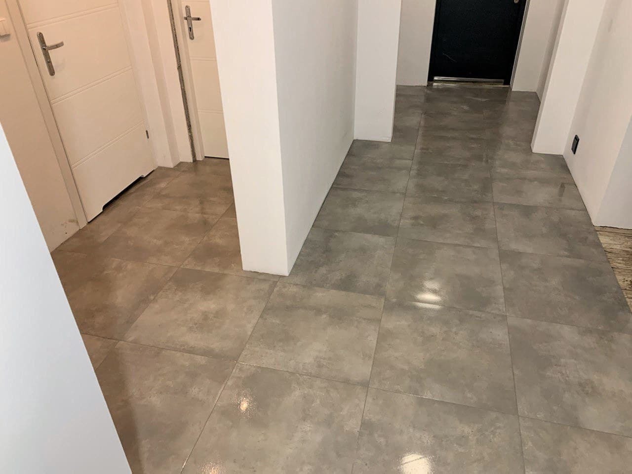 Ułożona szara, matowa terakota w korytarzu z białymi ścianami i drzwiami, widoczne fugi, odbicie światła na powierzchni płytek.