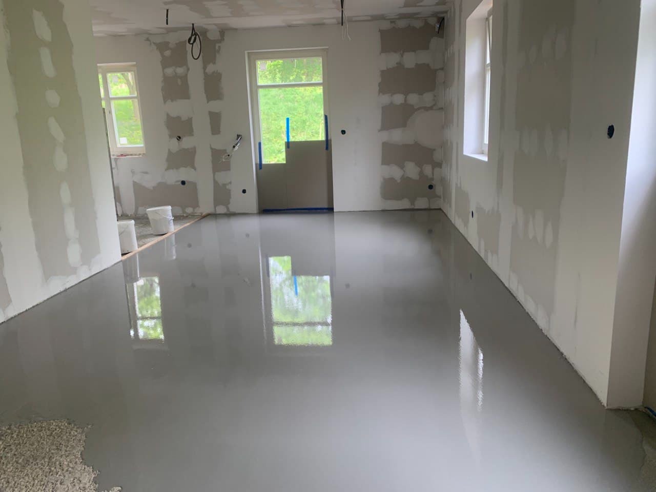 Wnętrze w trakcie prac wykończeniowych: świeżo wylana, gładka wylewka podłogowa, ściany częściowo pokryte gładzią gipsową, widoczne okna i drzwi zabezpieczone folią.