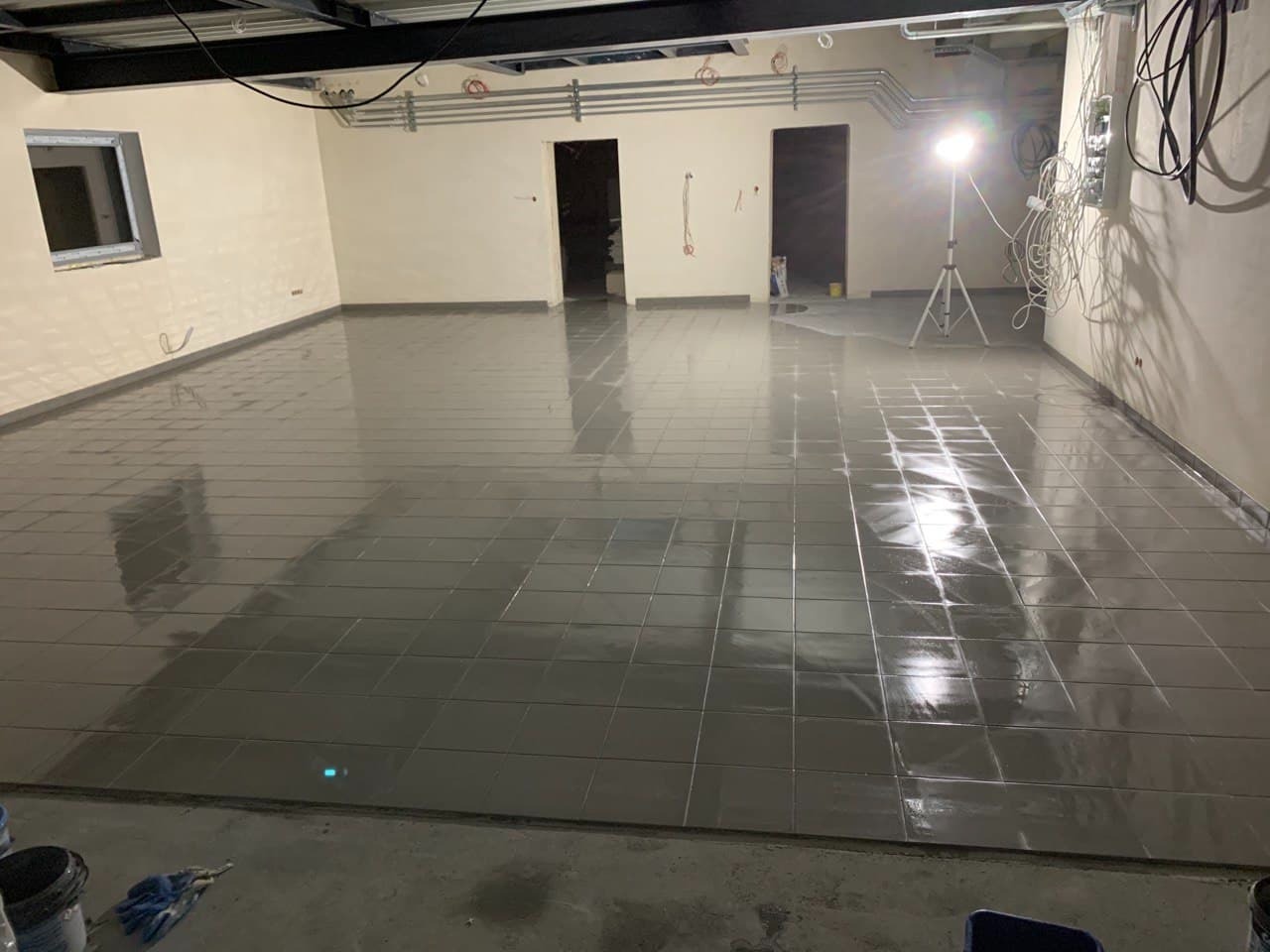 Świeżo położone, błyszczące, szare płytki podłogowe w pustym pomieszczeniu z otworami drzwiowymi i oknem, oświetlone lampą na statywie.