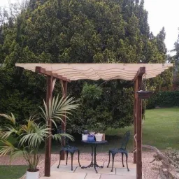 Drewniana pergola z falistym wykończeniem dachu, stojąca na wyłożonym kamieniem patio w ogrodzie, z palmą w doniczce po lewej stronie i małym stolikiem z krzesłami, na którym stoją doniczki...