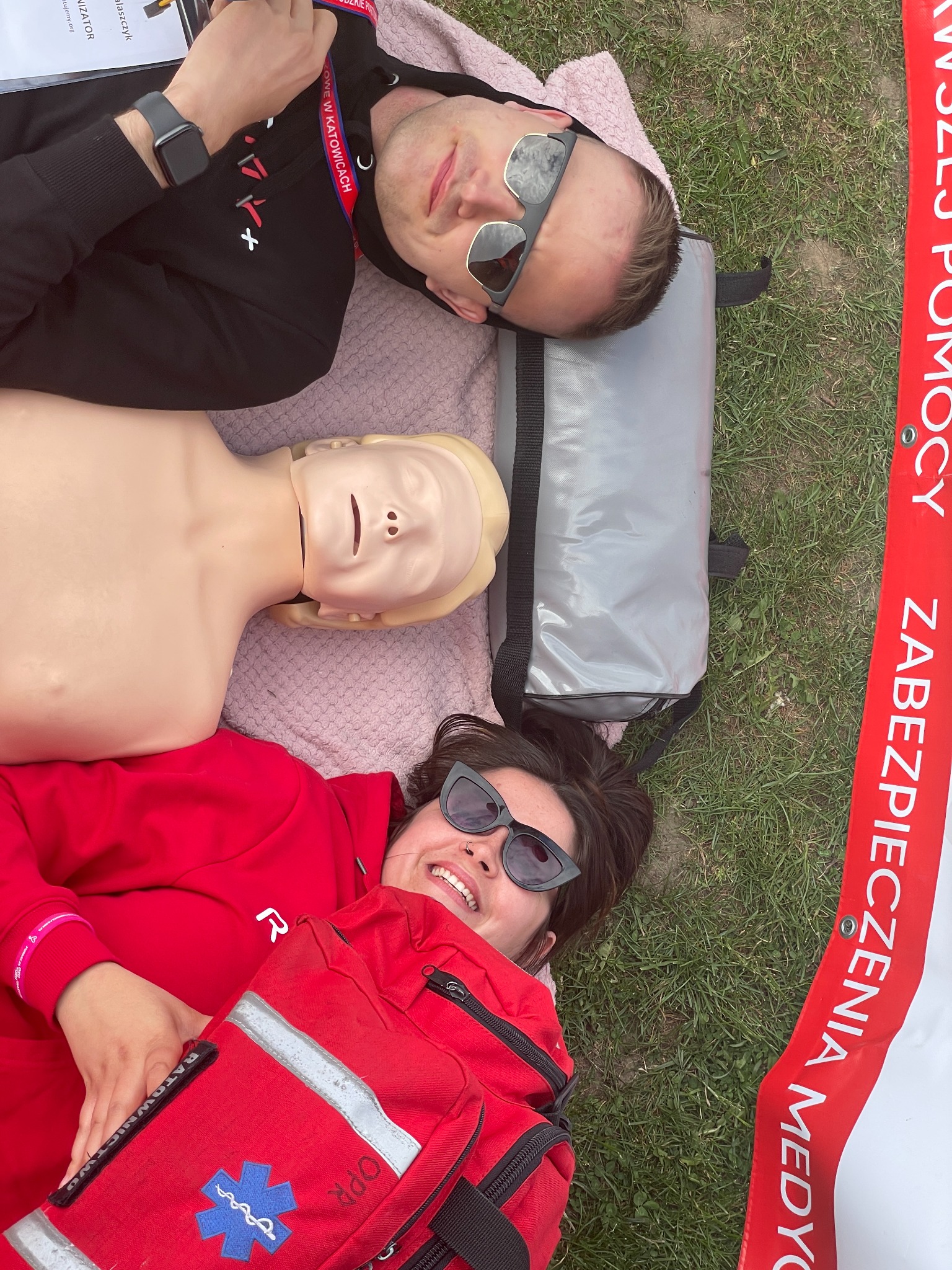 Instruktorzy pierwszej pomocy z manekinem do resuscytacji i torbą medyczną leżący na trawie podczas przerwy w szkoleniu.