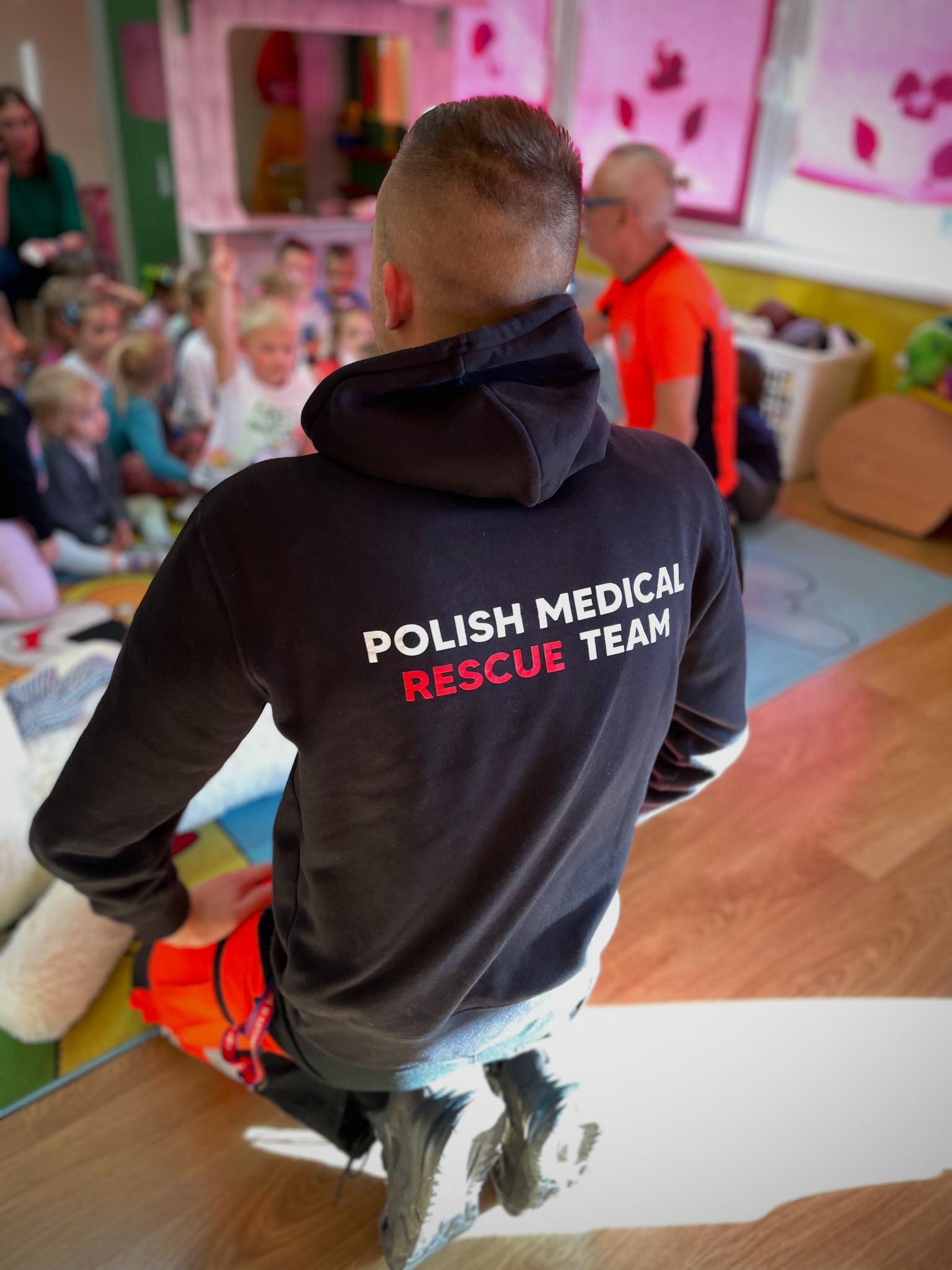 Instruktor w bluzie Polish Medical Rescue Team klęczy, demonstrując na fantomie resuscytację krążeniowo-oddechową podczas zajęć dla dzieci w przedszkolu.