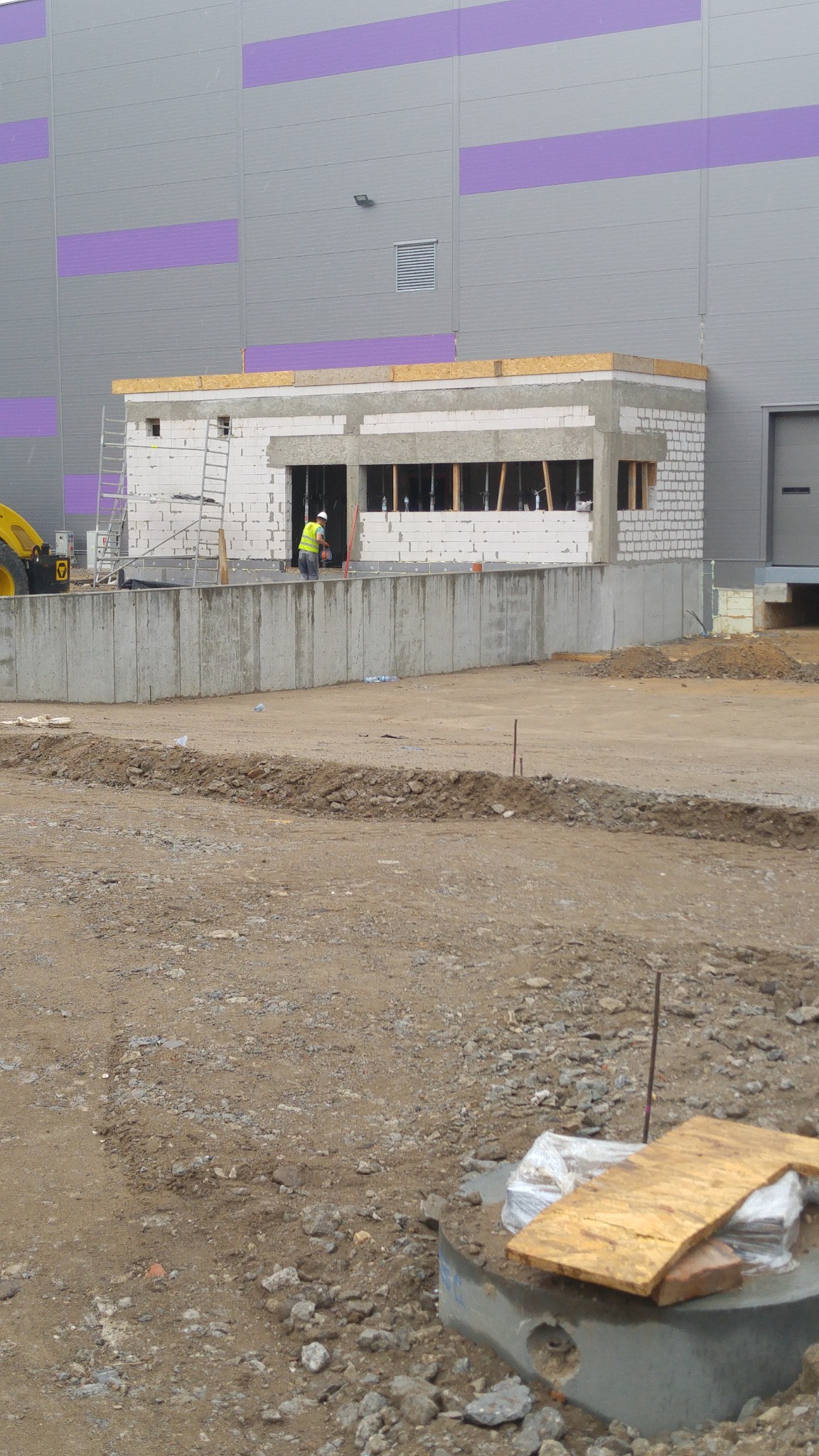 Budowa parterowego budynku z betonu komórkowego przy hali magazynowej, widoczny pracownik w kamizelce odblaskowej i kasku, rusztowanie oraz teren budowy