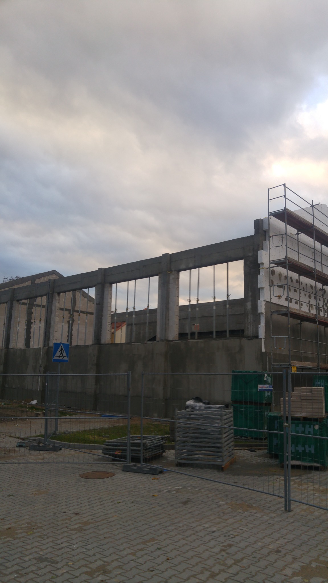 Surowy budynek żelbetowy w trakcie budowy, widoczne stalowe pręty zbrojeniowe, rusztowania i materiały izolacyjne, ogrodzenie placu budowy i znak ostrzegawczy.