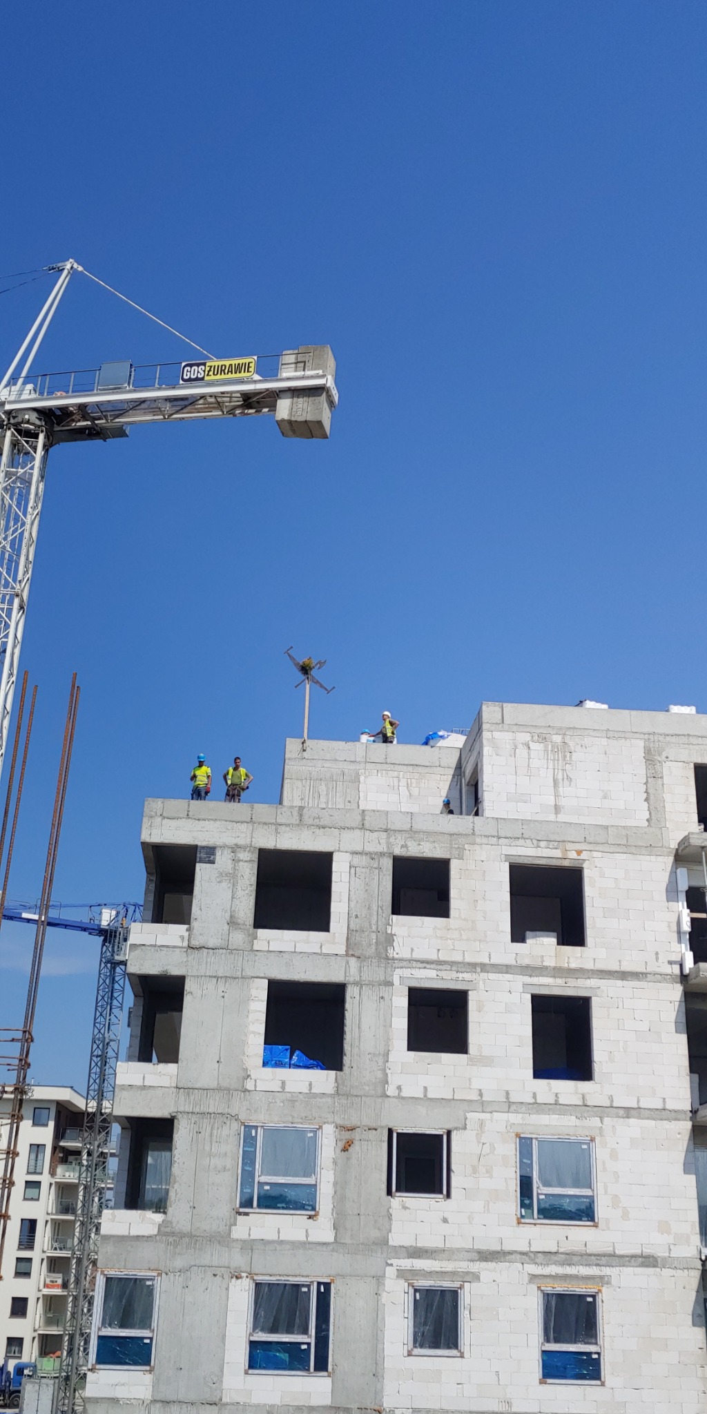 Budowa wielopiętrowego budynku z betonu komórkowego w trakcie realizacji, widoczni robotnicy w kaskach i kamizelkach odblaskowych na dachu, żuraw budowlany w tle, błękitne niebo.