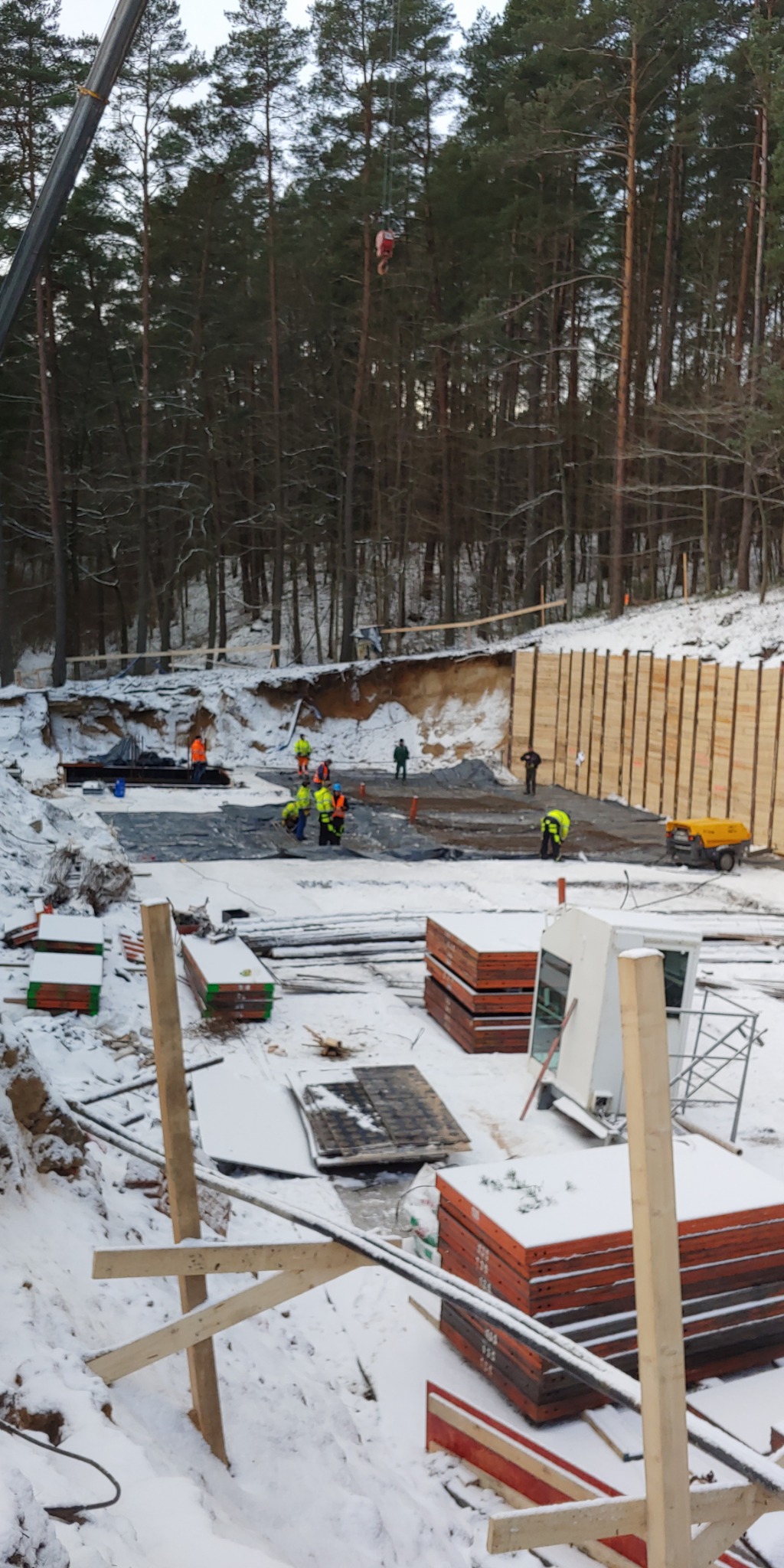 Budowa fundamentów w zimowej scenerii: pracownicy w odblaskowych kamizelkach przygotowują teren pod wylewkę, widoczne szalunki, zbrojenia i koparka w tle.