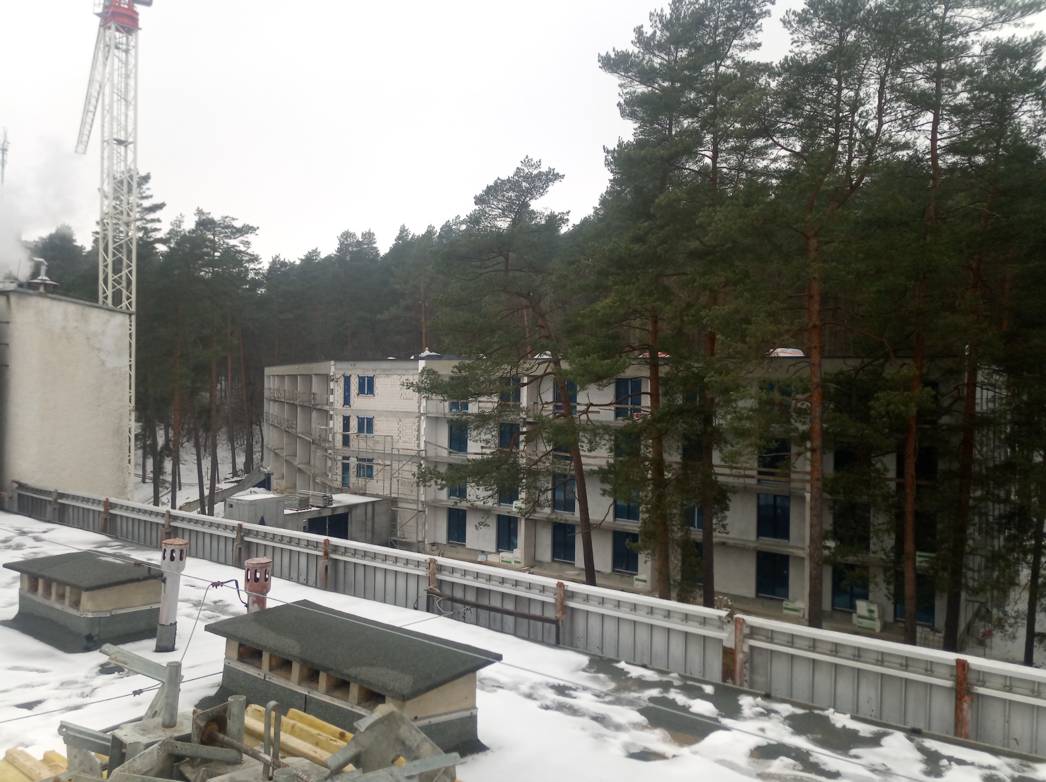 Wysoki budynek w stanie surowym otoczony lasem, widok z dachu innego budynku pokrytego śniegiem, w tle widoczny żuraw budowlany.