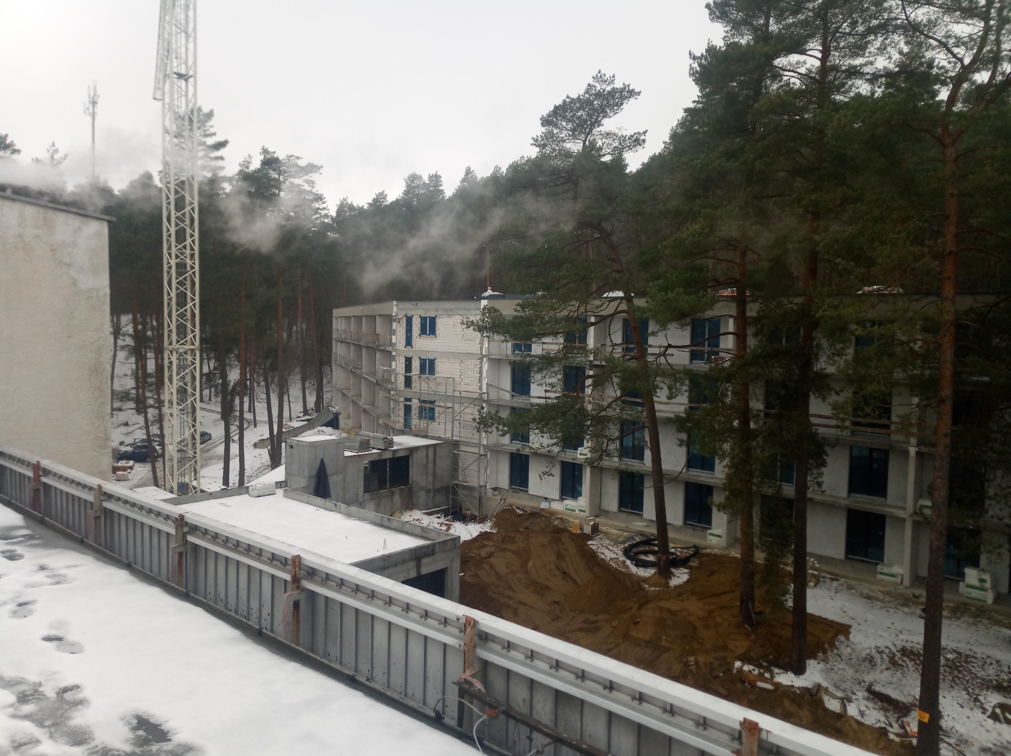 Wielopiętrowy budynek w trakcie budowy, otoczony lasem, widoczny dźwig i rusztowania, śnieg na pierwszym planie, z komina budynku wydobywa się para.
