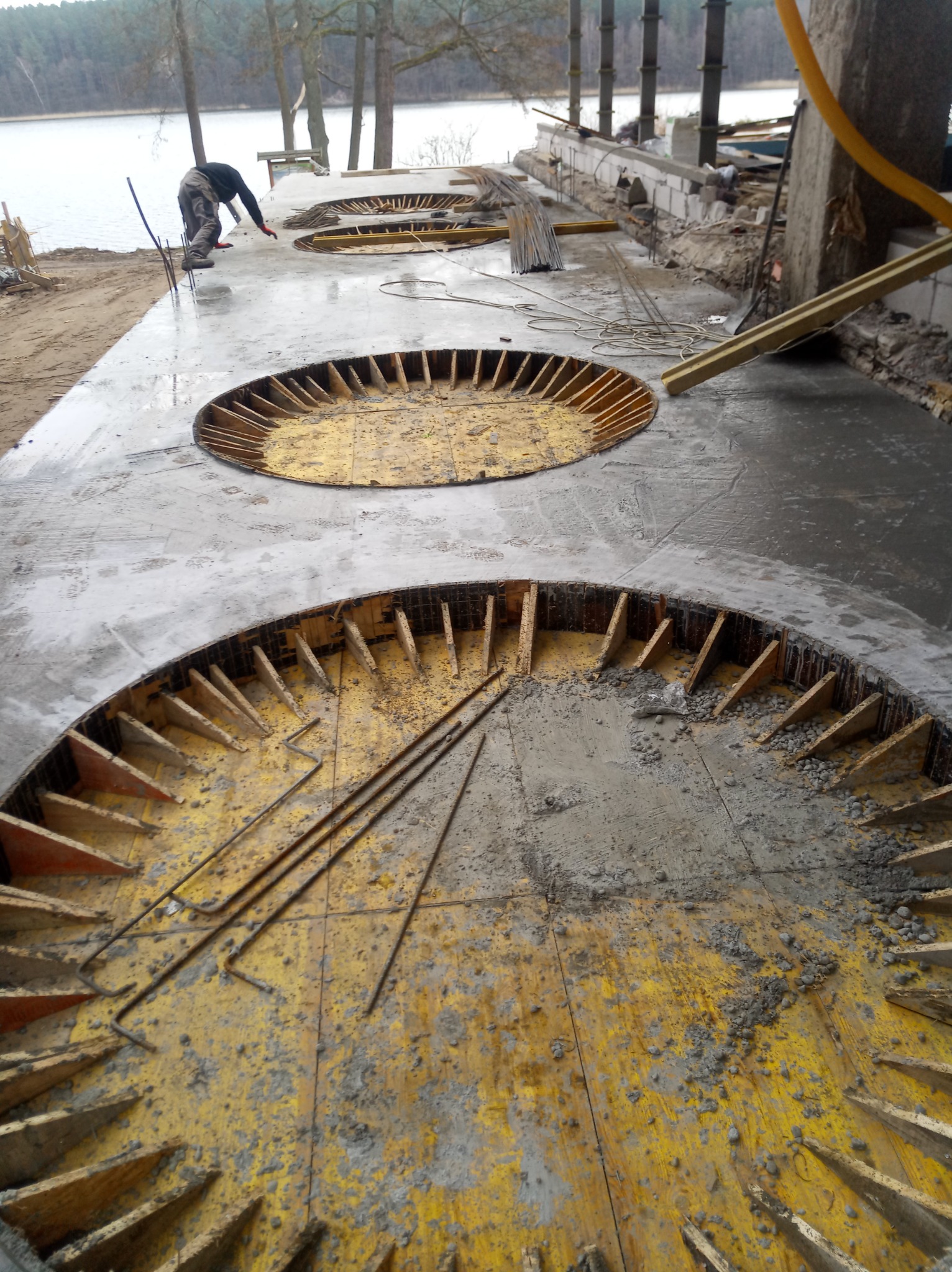 Wylewka betonowa z okrągłymi otworami z drewnianym szalunkiem na brzegu jeziora, widoczna osoba w oddali przy brzegu.