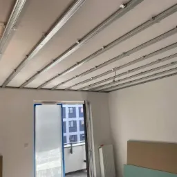 Montaż metalowego stelażu pod płytę kartonowo-gipsową na suficie w pomieszczeniu z wyjściem na balkon, widoczne profile, uchwyty montażowe i częściowo zamontowane płyty.