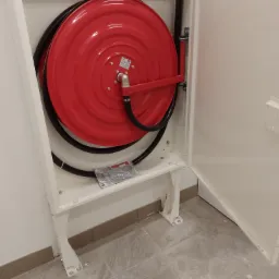 Czerwona zwijana szpula z wężem gaśniczym w białej szafce, widoczny zawór i zestaw montażowy, numer seryjny na drzwiczkach.