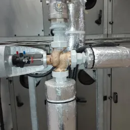 Złożony węzeł rur z zaworami i siłownikiem, izolowany folią aluminiową, na tle metalowej obudowy systemu wentylacyjnego.