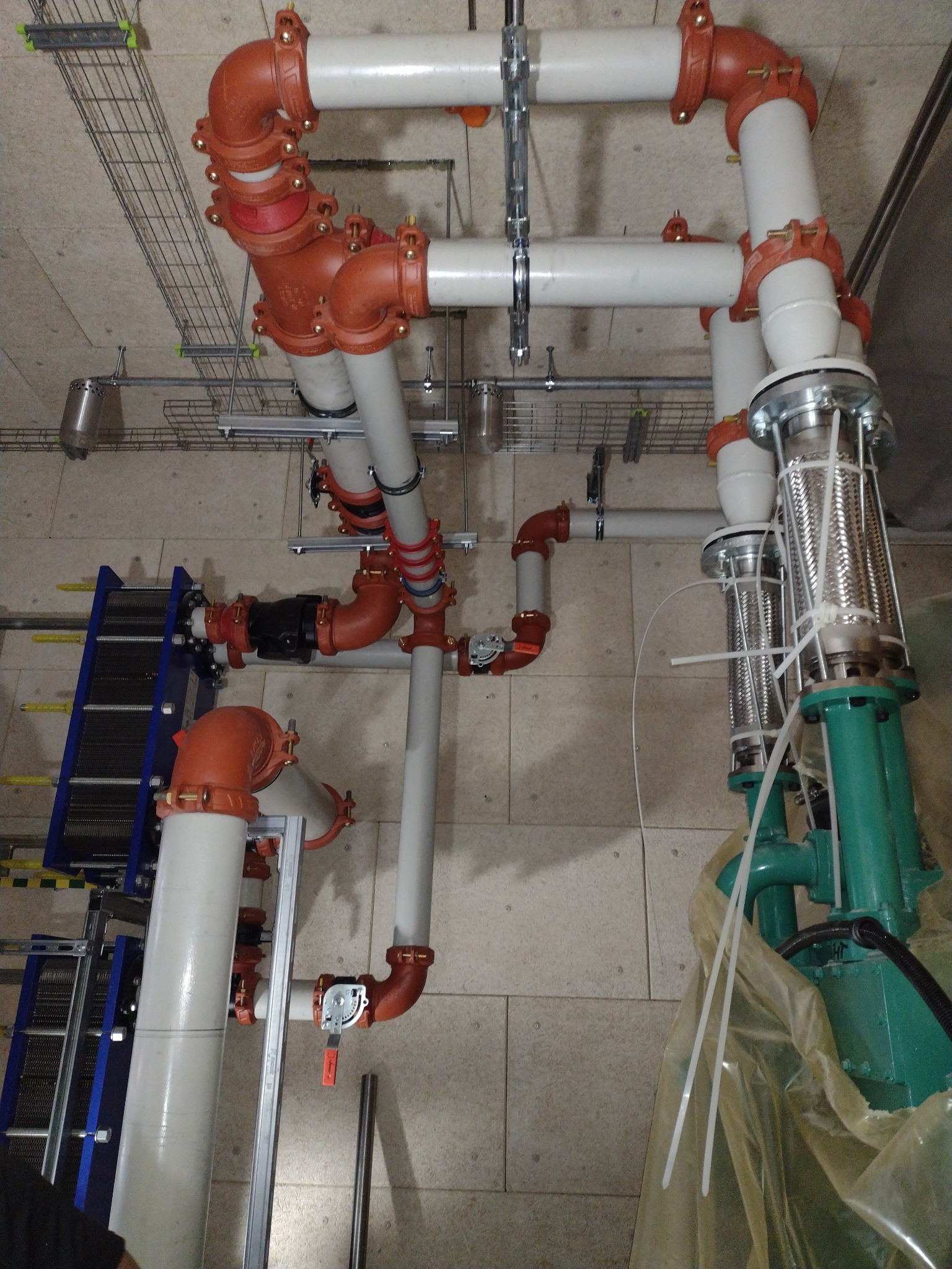 Złożona instalacja rur grzewczych i hydraulicznych z elementami w kolorze pomarańczowym i białym, widoczne zawory, metalowe kable i elementy mocujące pod sufitem.