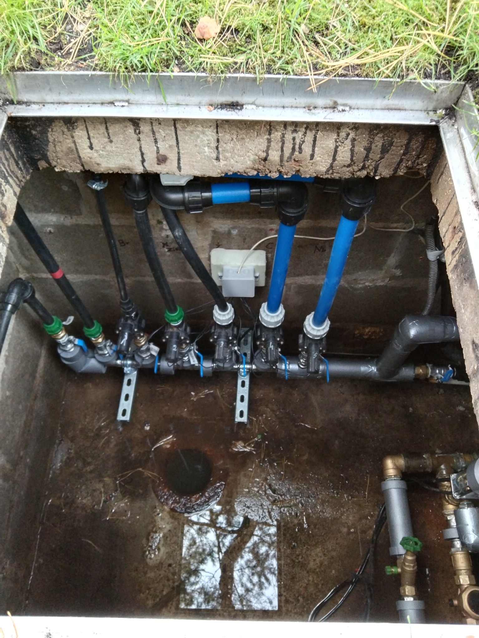 Studzienka rewizyjna z instalacją wodną, widoczne rury czarne i niebieskie, zawory, metalowe wsporniki, woda na dnie studzienki.