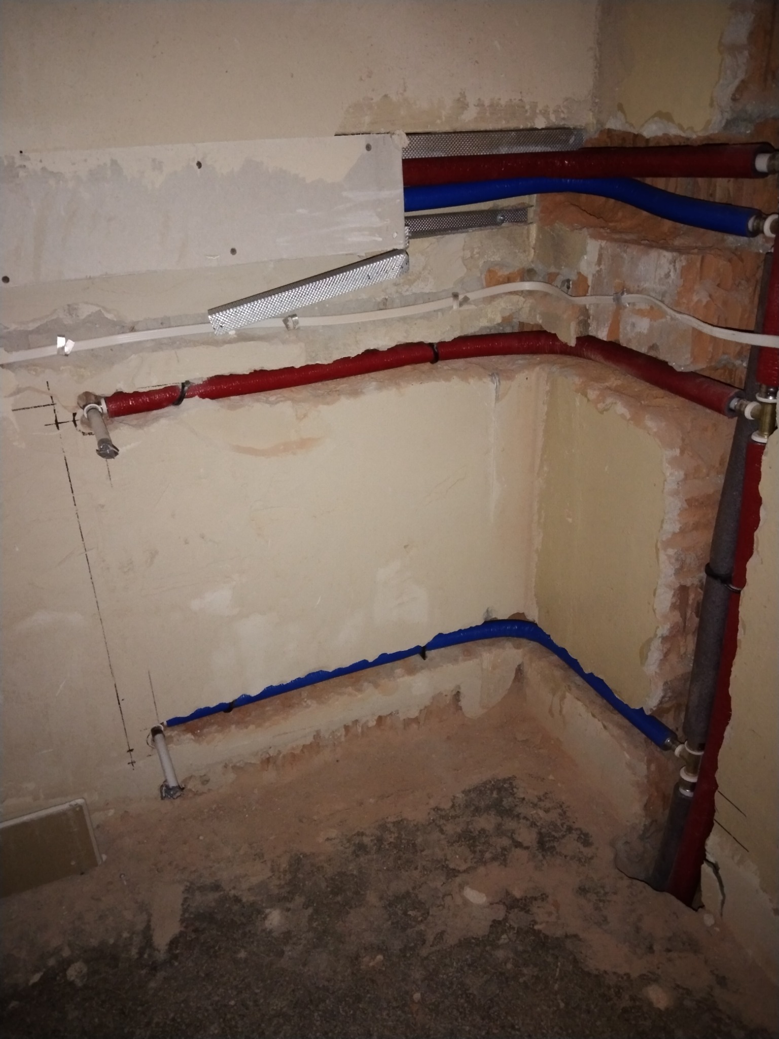 Surowe wnętrze w trakcie prac hydraulicznych: widoczne czerwone i niebieskie rury instalacyjne prowadzone wzdłuż ścian z fragmentami odsłoniętej cegły i niedokończonym tynkiem.