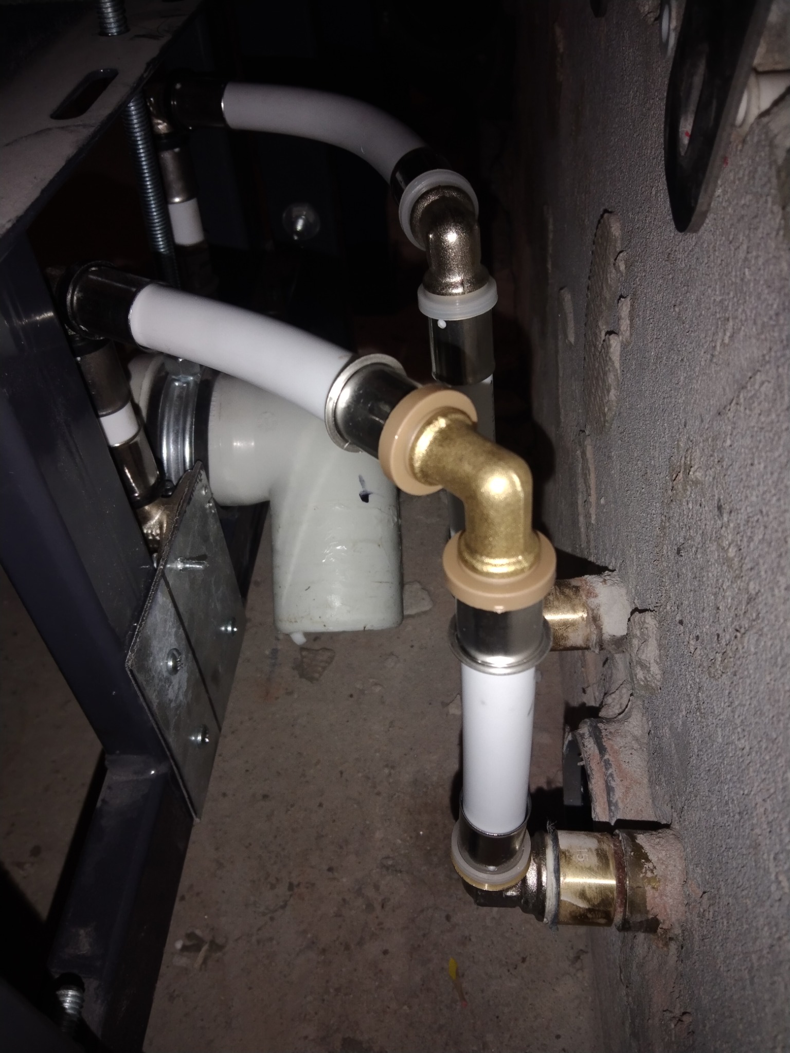 Fragment instalacji wodnej z białych rur i mosiężnych złączek, widoczne połączenie z szarą ścianą i element konstrukcyjny z metalu.