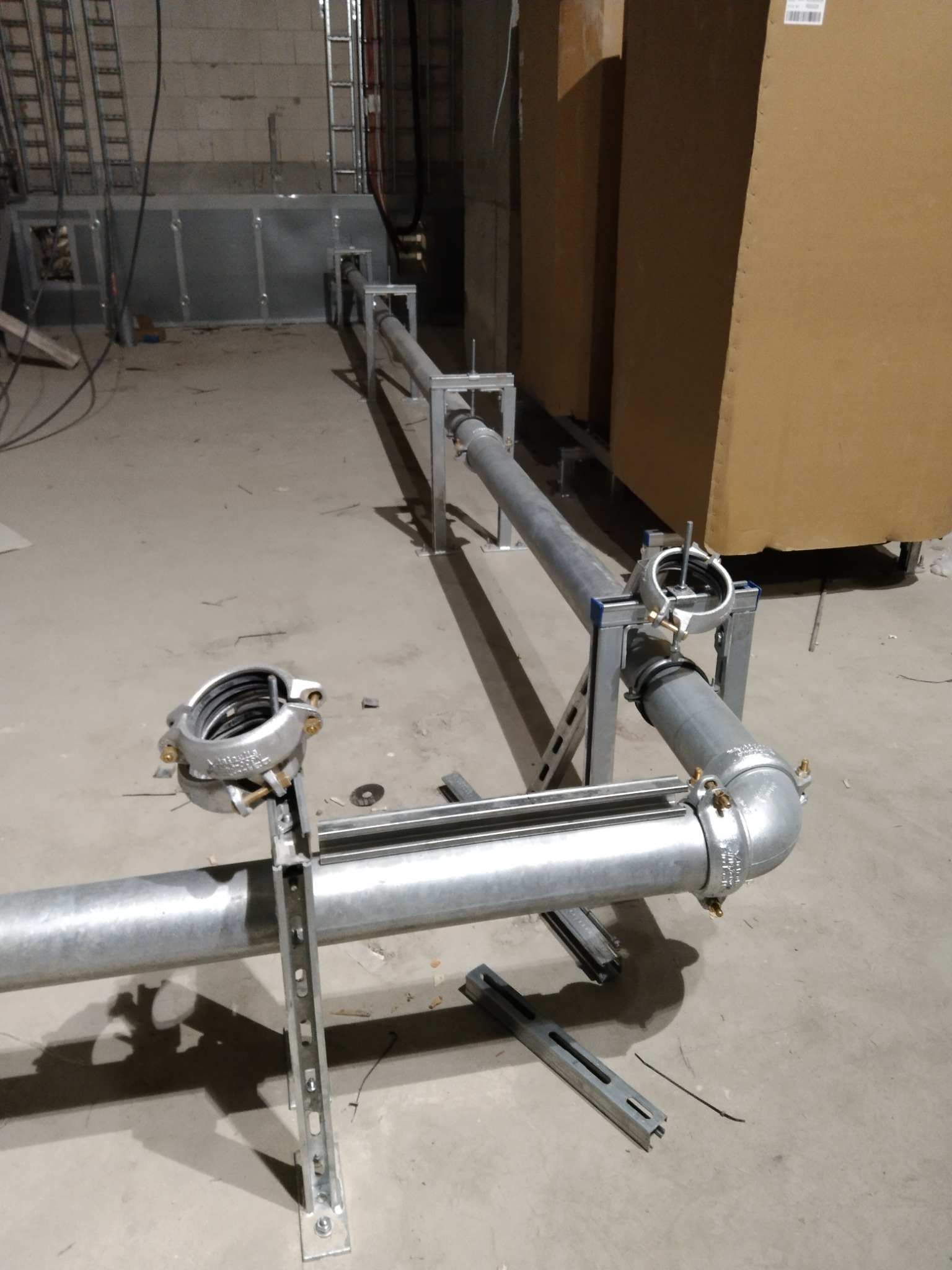 Szare rury instalacyjne, połączone złączkami i wspornikami, prowadzone wzdłuż ściany w surowym pomieszczeniu z widocznymi elementami konstrukcyjnymi i przewodami.